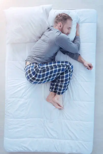 Cozy sleep. Top view on a young man wearing pajamas embracing his pillow while sleeping tight in a fetal position at home.