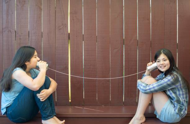 A 13-year old Eurasian girl is communicating with an 11-year old Eurasian girl using an old fashioned string telephone