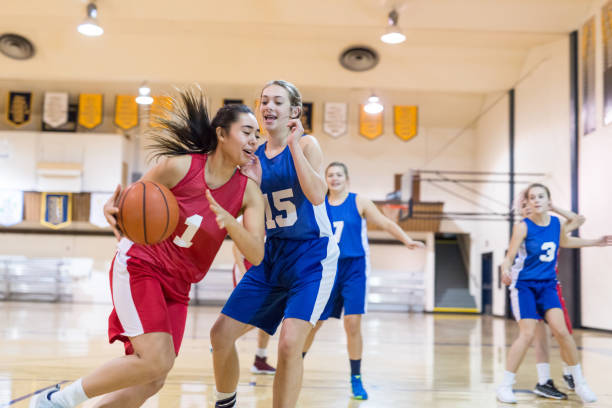 Girls high school basketball game An ethnic female player dribbles through the lane and tries to score. A defensive player is stepping up and trying to get there in time to prevent her from scoring. There are pennants and banners in the background. youth basketball indoor stock pictures, royalty-free photos & images
