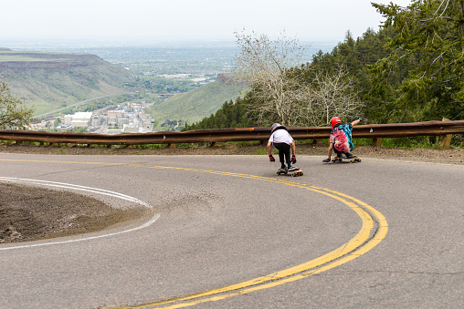 Golden, Colorado, USA - May 9, 2018: Two skateboarders cruising down a steep and winding mountain road at Lookout Mountain, with Table Mountains and city of Golden in background below.
