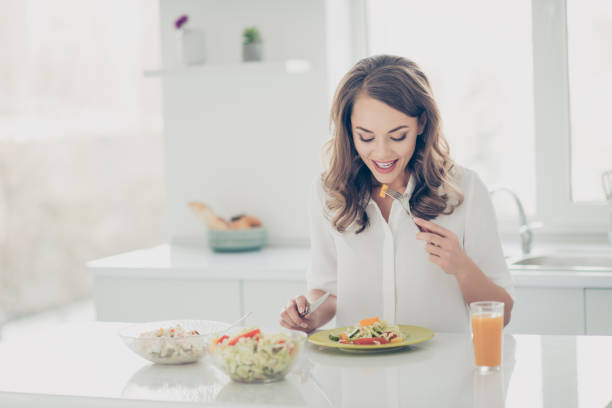 ritratto di donna carina, affascinante, elegante, gioiosa, allegra in camicia che mangia insalata di fitness con coltello e forchetta mettendo verdure in bocca mantenendo il programma di perdita di peso, dieta, stile di vita sano - breakfast salad leaf vegetable foto e immagini stock