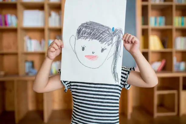 Photo of Young girl holding a self drawn portrait in front of her face