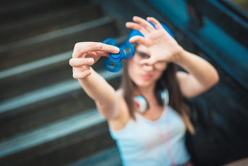 Playing with fidget spinner. Attractive young Caucasian woman wearing white shirt, denim shorts and blue baseball cap in street style is playing with a fidget spinner outdoors.