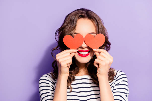 ritratto di bella ragazza creativa con denti bianchi rosso pomade che copre gli occhi di chiusura con due carta di cartone piccole figure del cuore amano i segni isolati su sfondo viola - hair care immagine foto e immagini stock