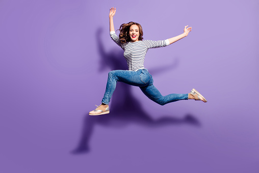 Portrait of crazy foolish girl jumping over in the air looking at camera having good stretching isolated on violet background, people life energy lifestyle concept