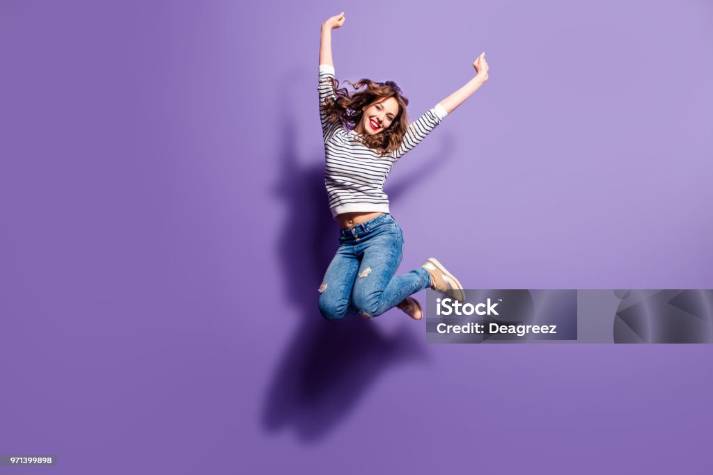 Ritratto di allegra ragazza positiva che salta in aria con pugni alzati guardando la telecamera isolata su sfondo viola. Concetto energetico delle persone della vita - Foto stock royalty-free di Donne