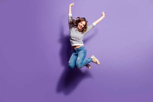 Photo of Portrait of cheerful positive girl jumping in the air with raised fists looking at camera isolated on violet background. Life people energy concept