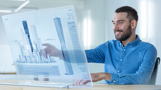 In the Futuristic Office Male Architects Works on the Transparent Computer Display with Skyscraper Building Model. Technologically Advanced Office Professional People Use 3D Modeling Software Application.