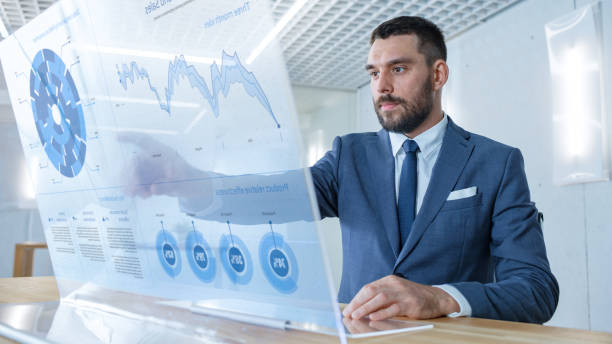 istock In the Near Future Successful Businessman Works on Computer with Transparent Display that Shows Stock Market Ticker Data, Numbers, Rises and Downs. 971403582