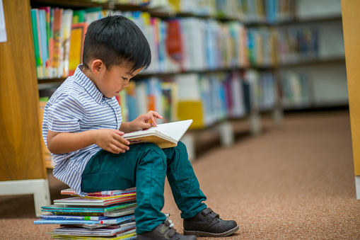 A young Asian boy is indoors in his elementary school library. He is reading a storybook while sitting on a stack of books.