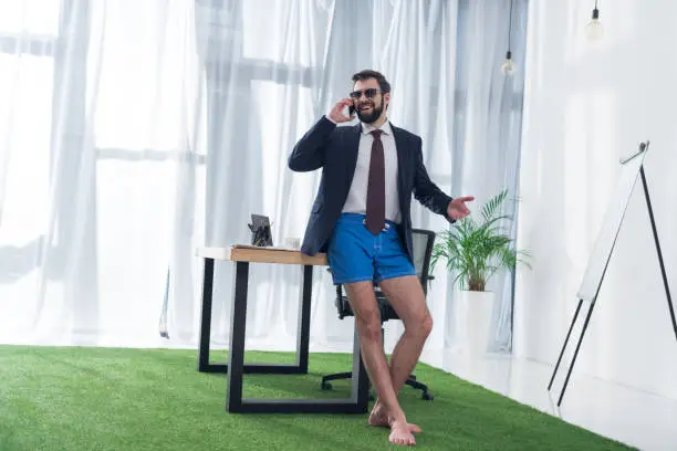 Photo of businessman in jacket and shorts talking on smartphone at workplace in office