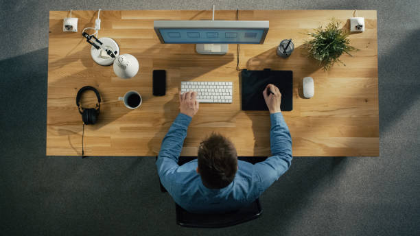 istock Top View of a Young Graphic Designer Working on a Digital Drawing Tablet Sitting at His Table. Also there is Desktop Computer, Headphones and Smartphone on the Table. 971549190