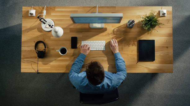 istock Top View of a Software Engineer Working on a Desktop Computer Sitting at His Table. Table is Neat, Smartphone, Plant, Coffee Cup, Headphones are Lying on it. 971549194