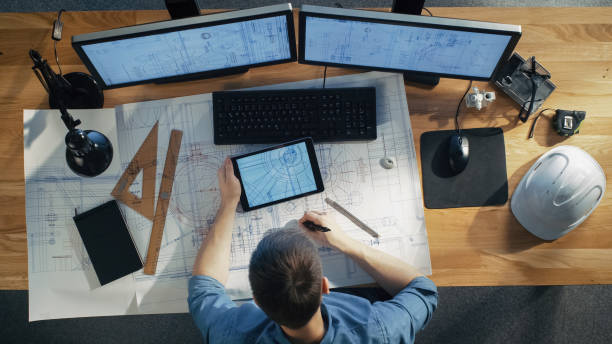 istock Top View of architectural Engineer Draws on His Blueprints, Compares with Tablet Computer with Green Screen, Using Desktop Computer Also. His Desk is Full of Useful Objects and Evening Sun. 971549300