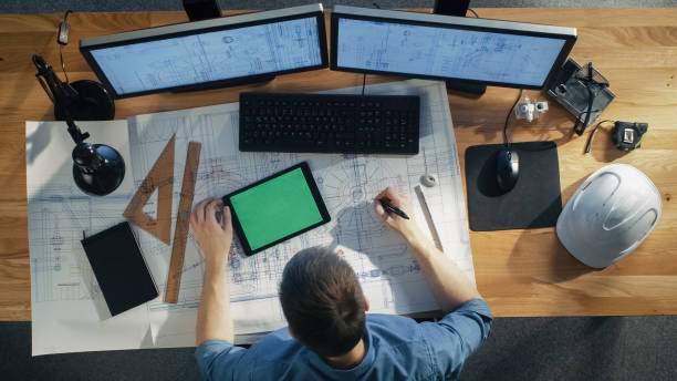 istock Top View of Architectural Engineer Draws on His Blueprints, Compares with Tablet Computer with Green Screen, Using Desktop Computer Also. His Desk is Full of Useful Objects and Evening Sun. 971549304