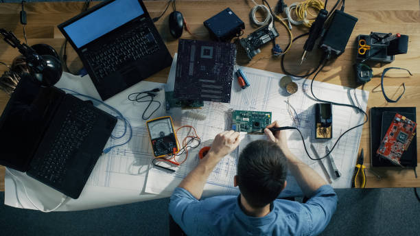 istock Top View of Young IT Technician Soldering Circuit Board. His Table is Full With Various Computer Parts, Motherboards, Laptops, Cables. Morning Sun Illuminates His Desk. 971549320