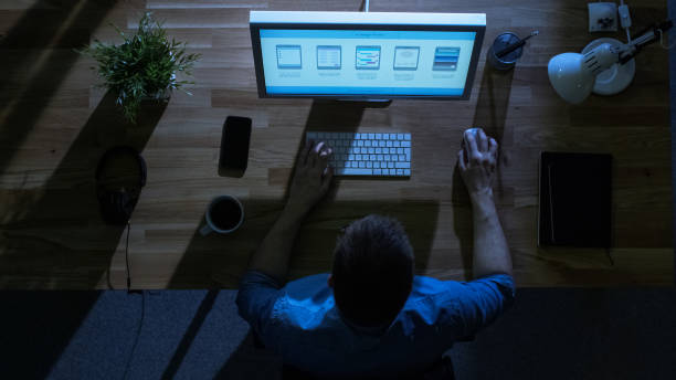 istock Top View of Young Creative Designer Working at His Desktop Computer at Night. His Table is Illuminated by Cold Blue Light From Outside. 971549338