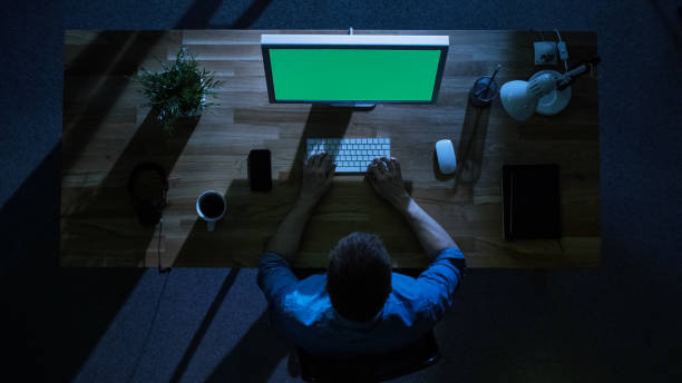 istock Top View of Male Programmer Working at His Desktop Computer with Mock-up Green Screen at Night. His Table is Illuminated by Cold Blue Light From Outside. 971549348