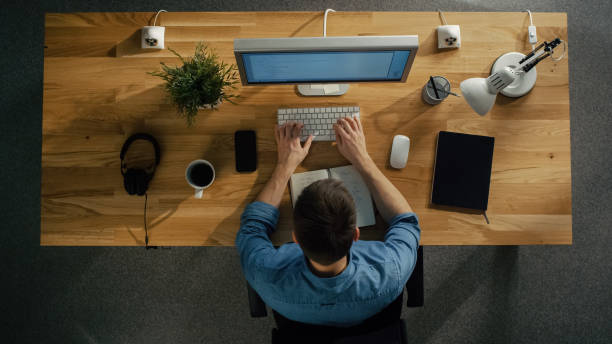 istock Top View of a Software Engineer Working on a Desktop Computer Sitting at His Table. Table is Neat, Smartphone, Plant, Coffee Cup, Headphones are Lying on it. 971549370