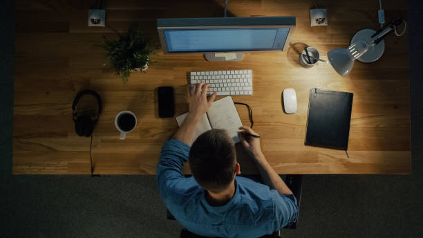 istock Top View of a Software Engineer Working on a Desktop Computer Sitting at His Table. Table is Neat, Smartphone, Plant, Coffee Cup, Headphones are Lying on it. 971549374