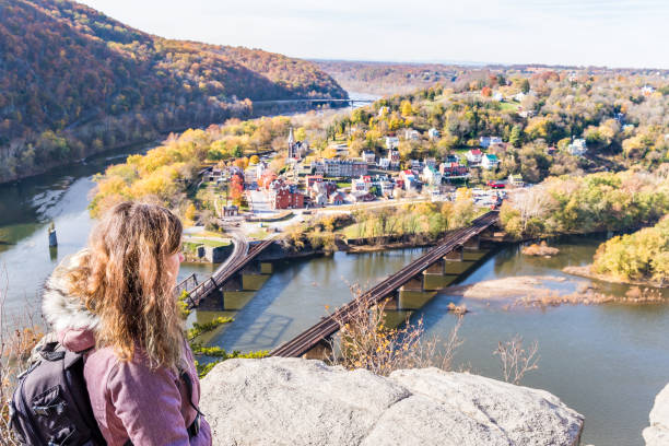 Hiker woman girl looking at cityscape overlook, colorful orange yellow foliage fall autumn forest with small village town by river in Harpers Ferry, West Virginia, WV Hiker woman girl looking at cityscape overlook, colorful orange yellow foliage fall autumn forest with small village town by river in Harpers Ferry, West Virginia, WV harpers ferry photos stock pictures, royalty-free photos & images