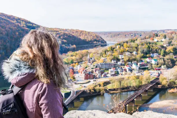 Overlook, hiker woman girl looking at cityscape, colorful orange yellow foliage fall autumn forest with small village town by river in Harpers Ferry, West Virginia, WV