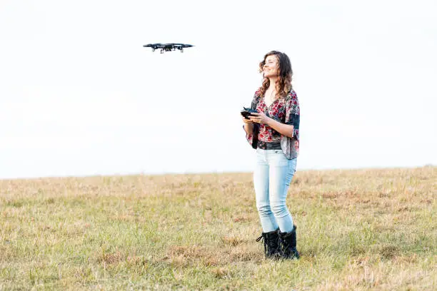 Photo of Young happy smiling woman controlling video camera drone in sky autumn or fall with boots in park in Virginia countryside open field hill