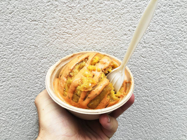 Hand holding a serving of Puerto Rican mofongo, fried plantains and garlic, with gourmet sauce in a sustainable paper street food bowl and a biodegradable fork