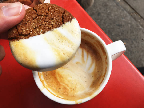 Hand holding sugar-crusted ginger cookie dipped in the foam of a cup of latte outdoors