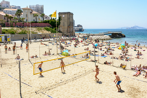 Marseille, France - May 19, 2018: Two mixed teams play beach volleyball on a court of the Catalans beach while people enjoy a warm and sunny summery day by the sea by sunbathing on the beach.
