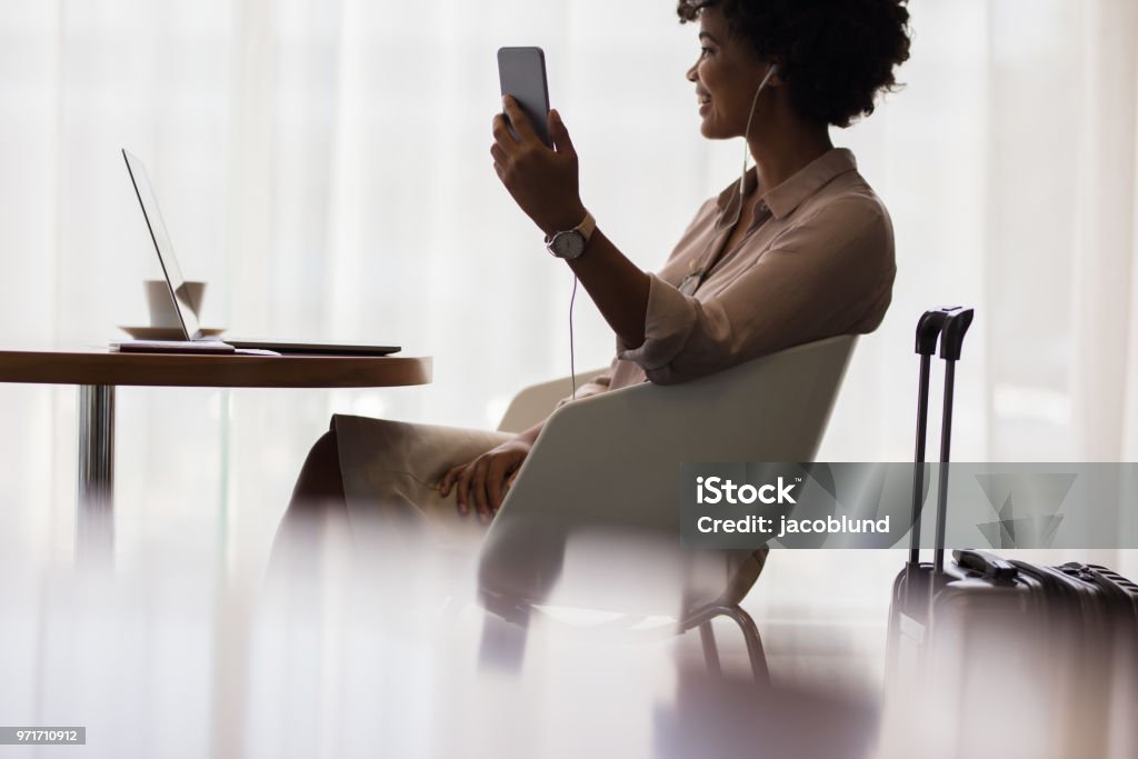 Woman at airport lounge making video call Happy woman making video call from mobile phone while sitting at airport lounge. Business woman waiting for flight at airport and using smart phone. Airport Departure Area Stock Photo