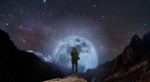 a man with backpack standing on mountain peak at night, and silhouette mountain with bright full moon and sky full of stars
