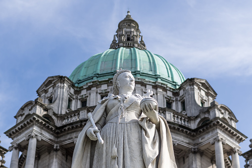 Belfast, Northern Ireland - June 5, 2018:  Statue of Queen Victoria which stands in front of Belfast City Hall.  \n\nThe statue was created by the sculptor Sir Thomas Brock to celebrate the Diamond Jubilee of Queen Victoria in 1897.  It was unveiled by her son, King Edward VII in 1903. Carved from Sicilian marble and standing 11 feet high, this memorial is accompanied on each side by life size bronze figures representing spinning and shipbuilding (two key industries in Belfast's growth) \n\nIn 1888 Queen Victoria granted Belfast the status of the city and it was agreed that a new city hall was required to reflect this new status. City Hall opened its doors on the first of August 1906, at a time of unprecedented prosperity and industrial might for the city. \n\nThe new City Hall was designed by Alfred Brumwell Thomas in the Baroque Revival style and constructed in Portland stone. The building cost £369,000 to complete, the equivalent around 128 million pounds today.