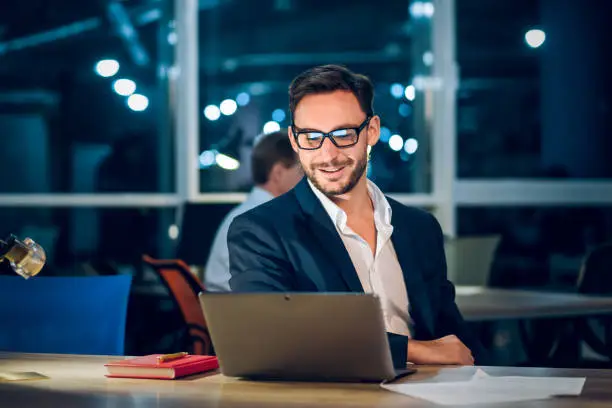 Attractive businessman smiling while working with laptop. Successful wealthy man with glasses and short beard working in office behind computer late in evening.