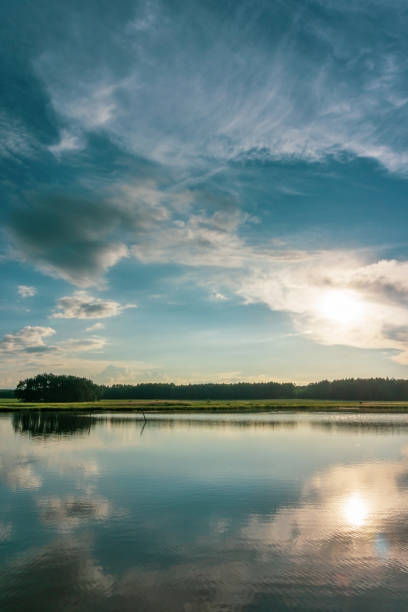 istock Vertical photo with summer landscape. Small pond is in foreground with reflected blue evening sky full of dramatic clouds. Green meadow with trees is in background. 971986708