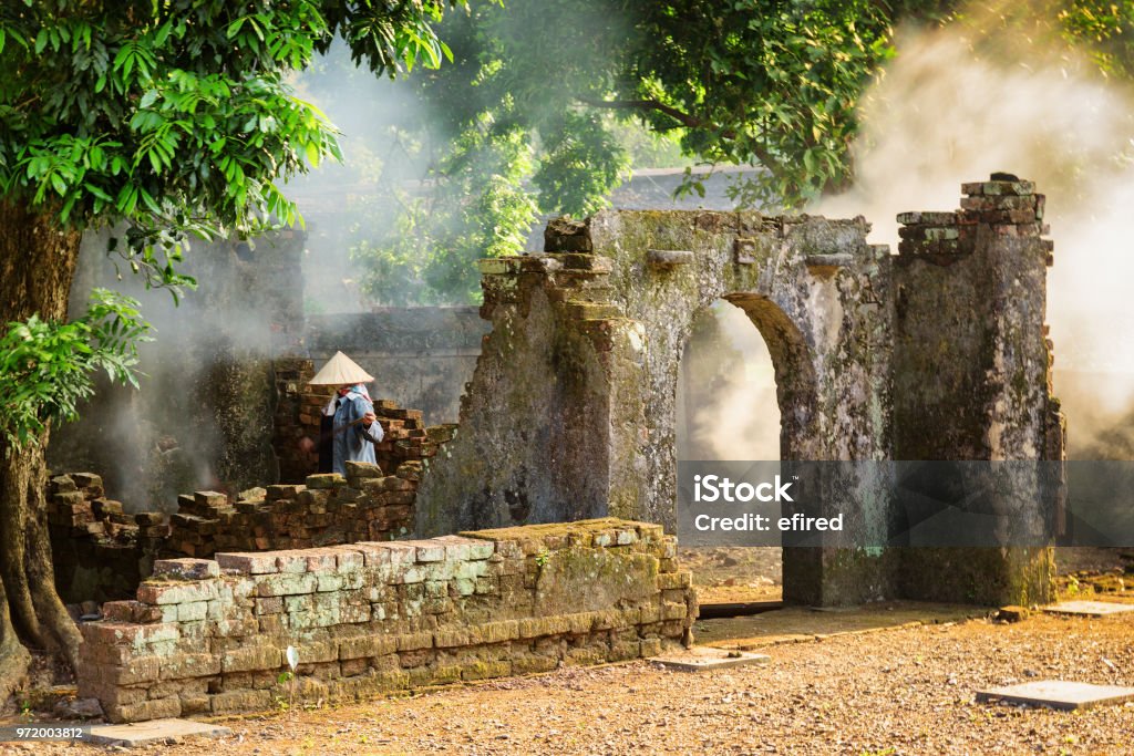 Vietnamese woman among ruins of old buildings, Hue, Vietnam Unknown Vietnamese woman in traditional bamboo hat among scenic ruins of old buildings at the Tu Duc Royal Tomb in Hue, Vietnam. Hue is a popular tourist destination of Asia. Huế Stock Photo