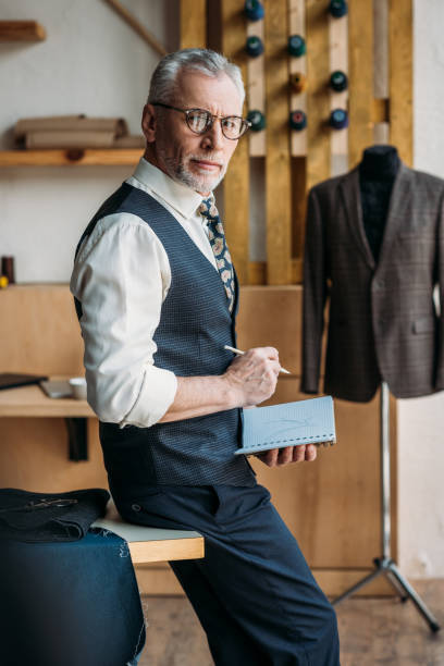 istock senior tailor holding notebook and pencil at sewing workshop and looking at camera 972010358