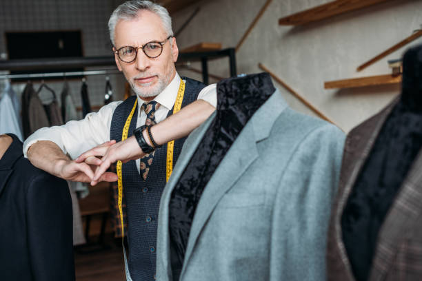 istock stylish mature tailor with measuring tape leaning on mannequin with jacket at sewing workshop 972013622