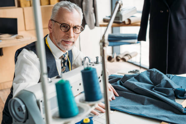 istock handsome tailor sewing cloth with sewing machine and looking at camera at sewing workshop 972017336
