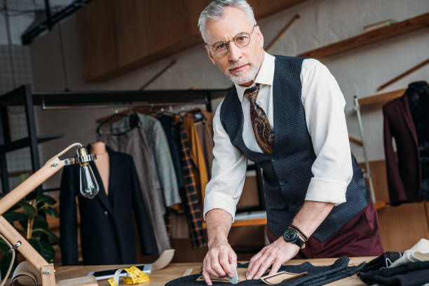 istock handsome mature tailor marking cloth pattern with chalk at sewing workshop and looking at camera 972035754