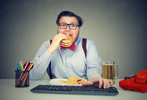 Overweight business man sitting at working place with computer and consuming fast food looking super hungry at