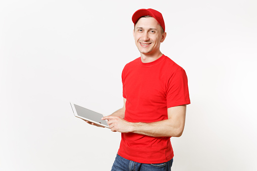 Delivery man in red uniform isolated on white background. Male in cap, t-shirt, jeans working as courier or dealer, holding tablet pc computer with blank empty screen. Copy space for advertisement