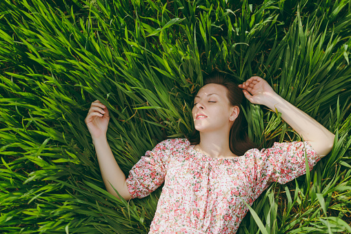 Young smiling tender beautiful woman with closed eyes in light patterned dress lying on grass resting in sunny weather in field on bright green background. Spring nature. Lifestyle, leisure concept