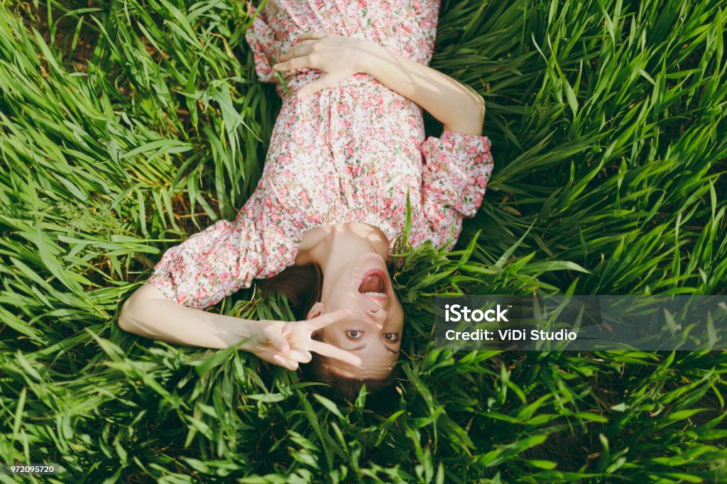 Joven Divertida Muy Contento A Hermosa Mujer En Vestido Estampado Ligero Acostado En El Pasto Mostrando Signo De Victoria Descansando En Tiempo Soleado En El Campo De Fondo Verde Brillante Naturaleza De Joven Divertida Muy Contento A Hermosa Mujer En Vestido Estampado Ligero Acostado En El Pasto Mostrando Signo De Victoria Descansando En Tiempo Soleado En El Campo De Fondo Verde Brillante Naturaleza De