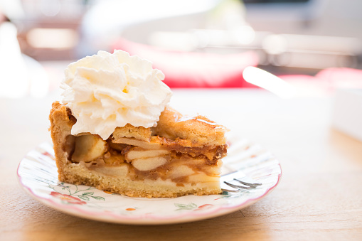 slice of Dutch apple pie with whipped cream on saucer, table, against blur background