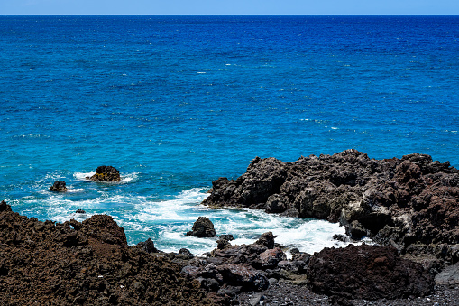 Bright blue Pacific Ocean with a rough black lava rock shoreline and crashing waves, Hawaii