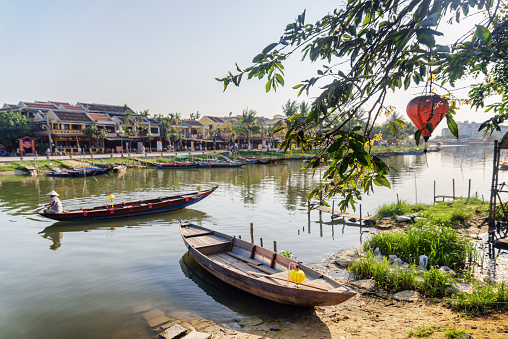 Beautiful view of wooden boat parked on bank of the Thu Bon River in Hoi An Ancient Town (Hoian), Vietnam. Vietnamese woman in traditional bamboo hat on boat is visible in background.