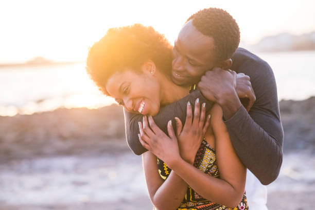 abraço e desfrutar de raça africana preta par de modelo homem e mulher jovem 25 anos de idade fica apaixonado e toca a brincar e brincar juntos. pôr do sol no fundo na praia com ondas e oceano. localização de tenerife - 25 30 years adult african descent bonding - fotografias e filmes do acervo