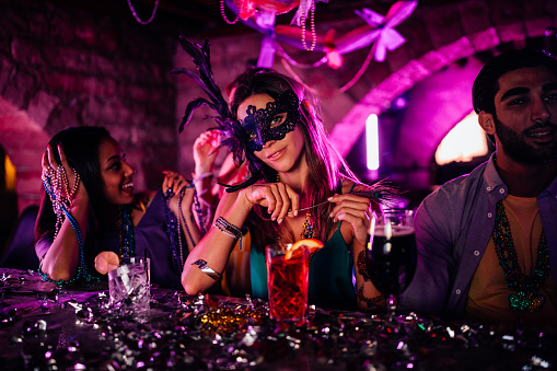 Young woman with mask sitting at bar counter, drinking cocktails and celebrating Mardi Gras
