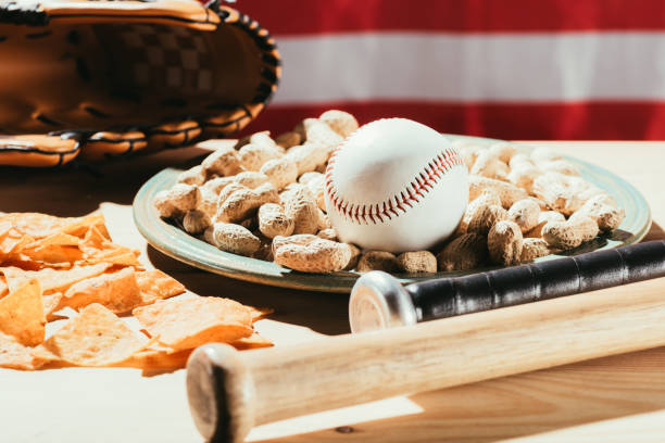 zbliżenie kijów baseballowych, piłka baseballowa na talerzu z orzeszkami ziemnymi, przekąski i skórzane rękawiczki na drewnianym stole z flagą usa za - baseball baseballs peanut american culture zdjęcia i obrazy z banku zdjęć